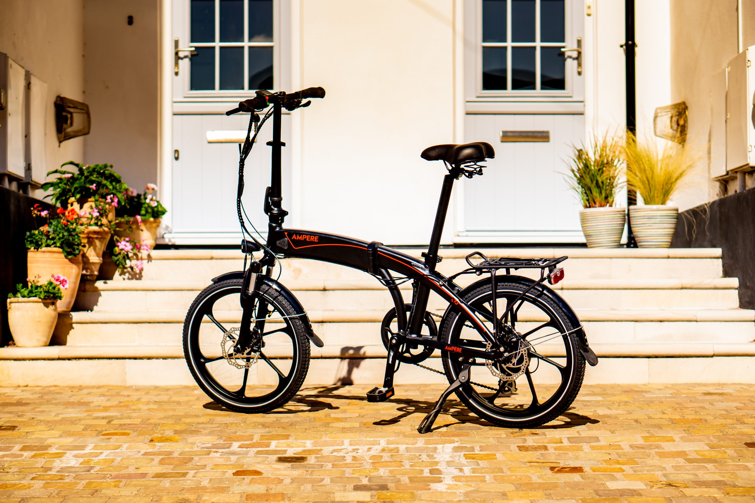 A black and red Ampere Electric folding bike in front of a scenic house