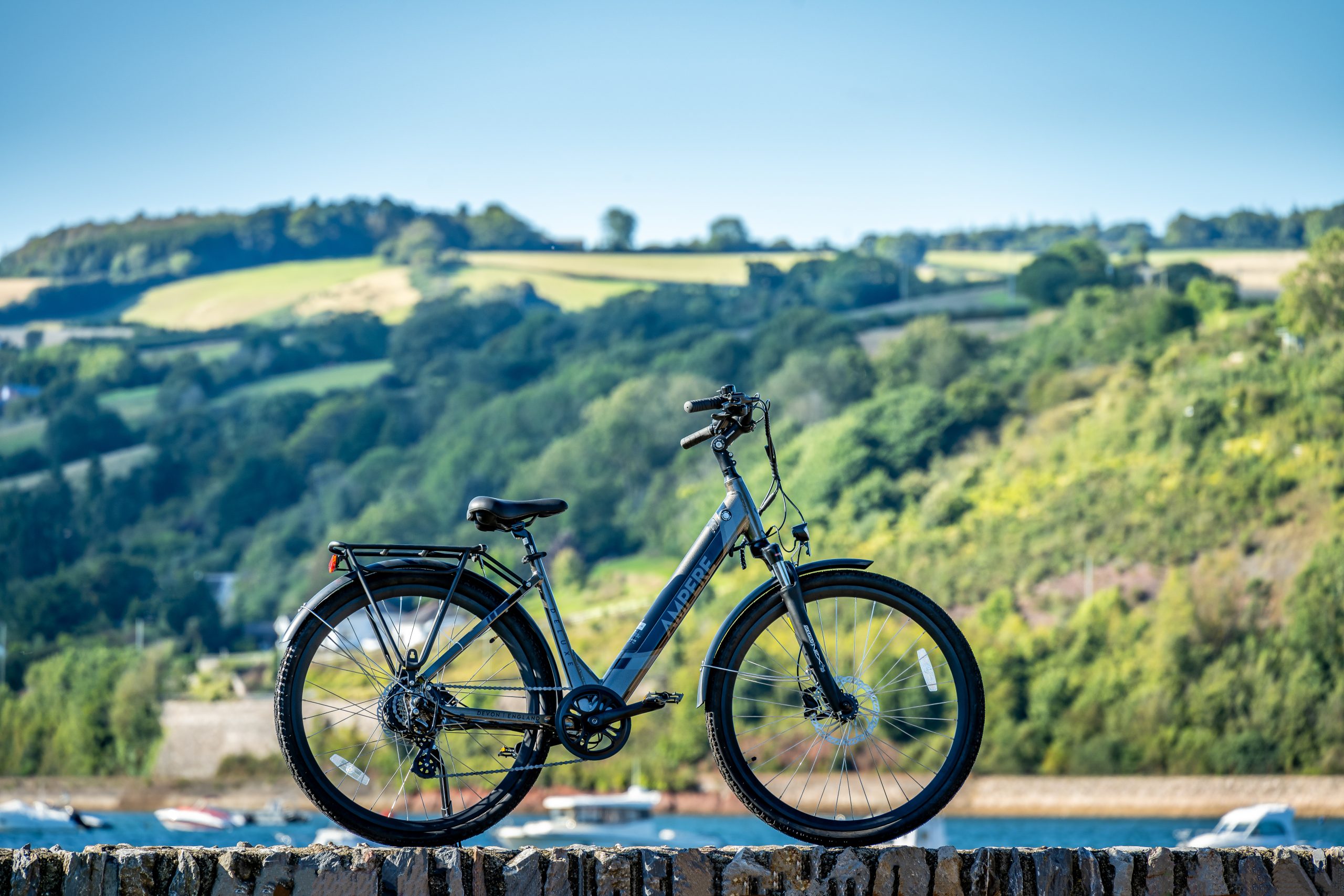 A grey Ampere Electric bike on a wall overlooking green hills and a harbour.